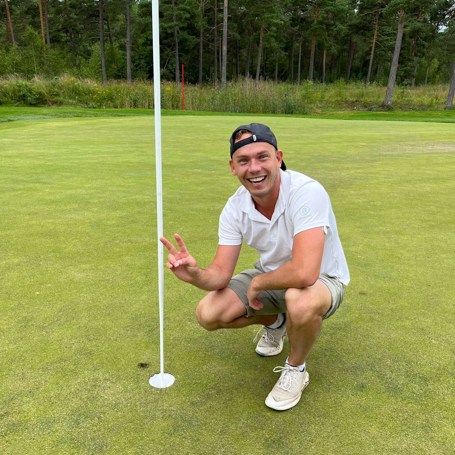 Smiling man in a white polo shirt and cap makes a victory sign next to a golf hole on a green golf course with forest in the background.