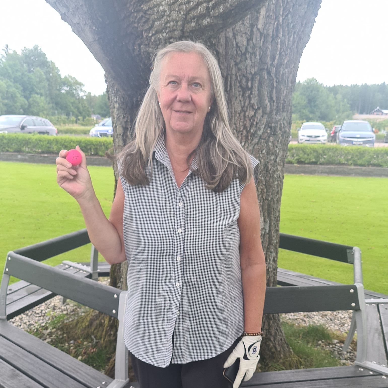 Kvinna med långt grått hår håller en rosa golfboll och står framför ett träd på en golfbana med bilar i bakgrunden.