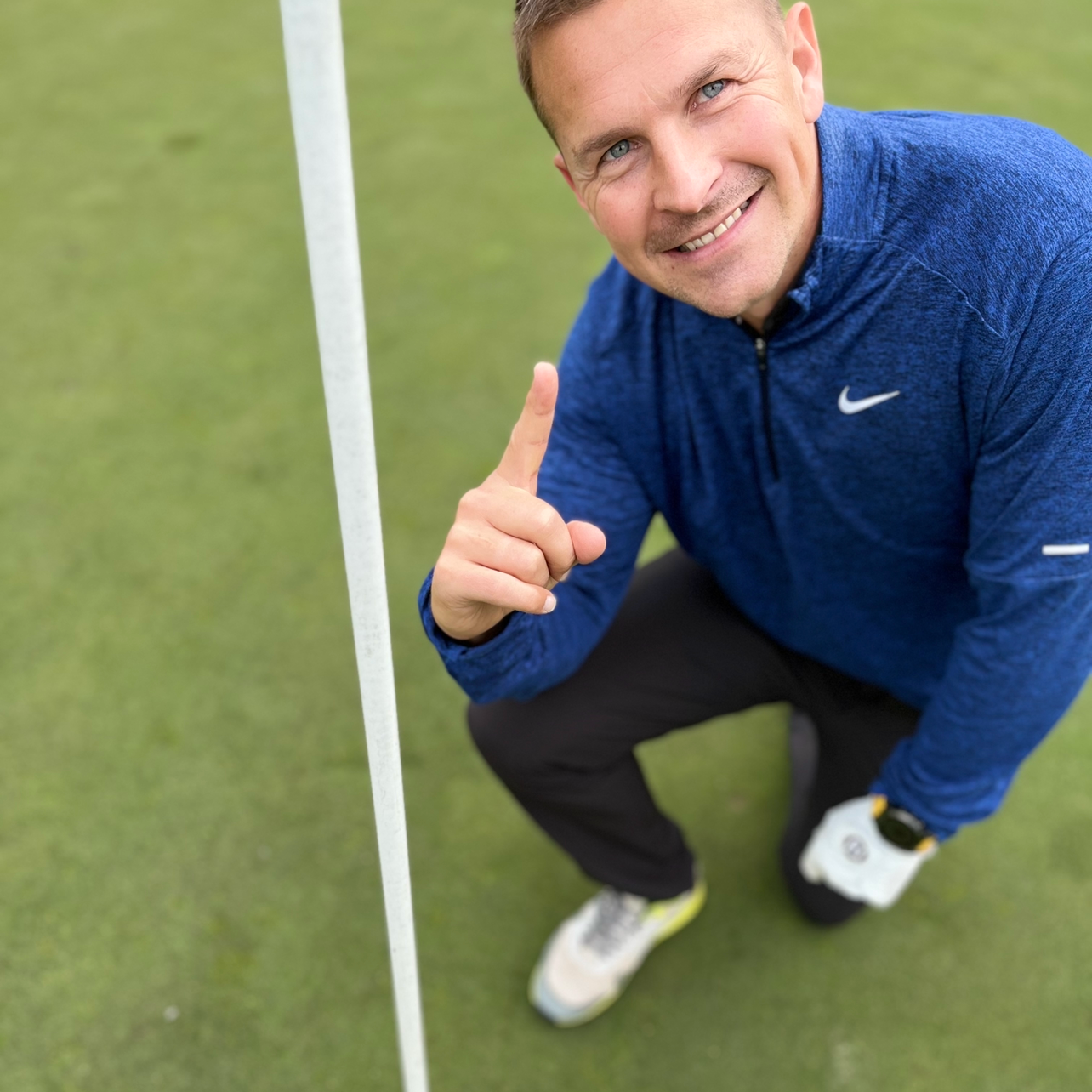 Mand i blå Nike-trøje knæler ved siden af et golf-flag, smiler og holder én finger op på en golfbane.