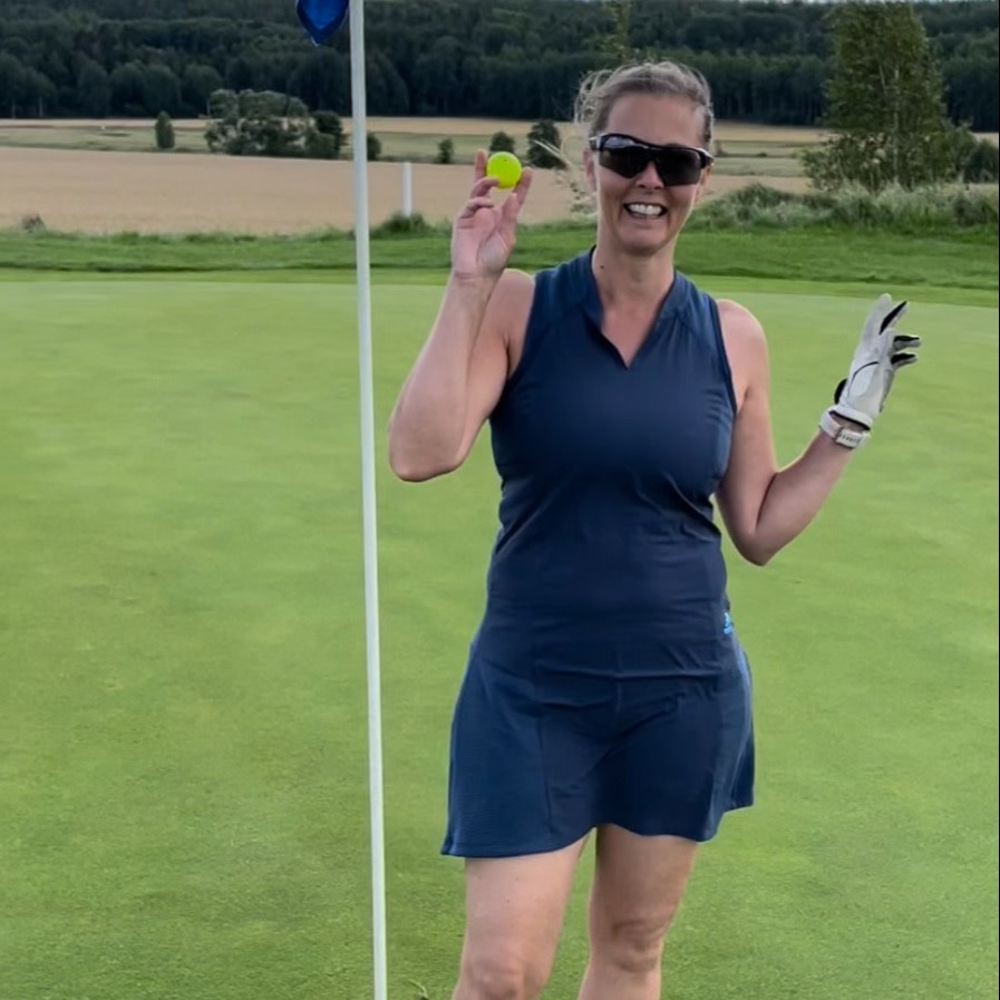 Kvinde i blå golfkjole og solbriller holder en golfbold op og smiler ved siden af en flagstang på en golfbane.