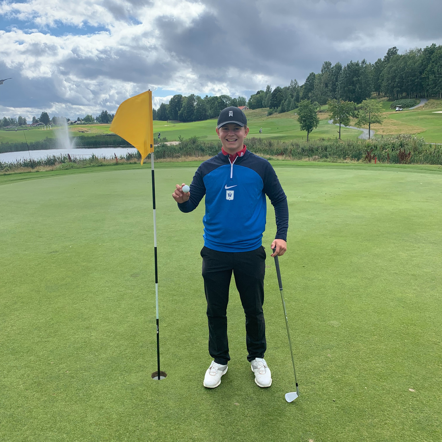 En golfspiller står på greenen ved siden af hullet og holder en golfbold op, med en golfkølle i den anden hånd og et gult flag i baggrunden.