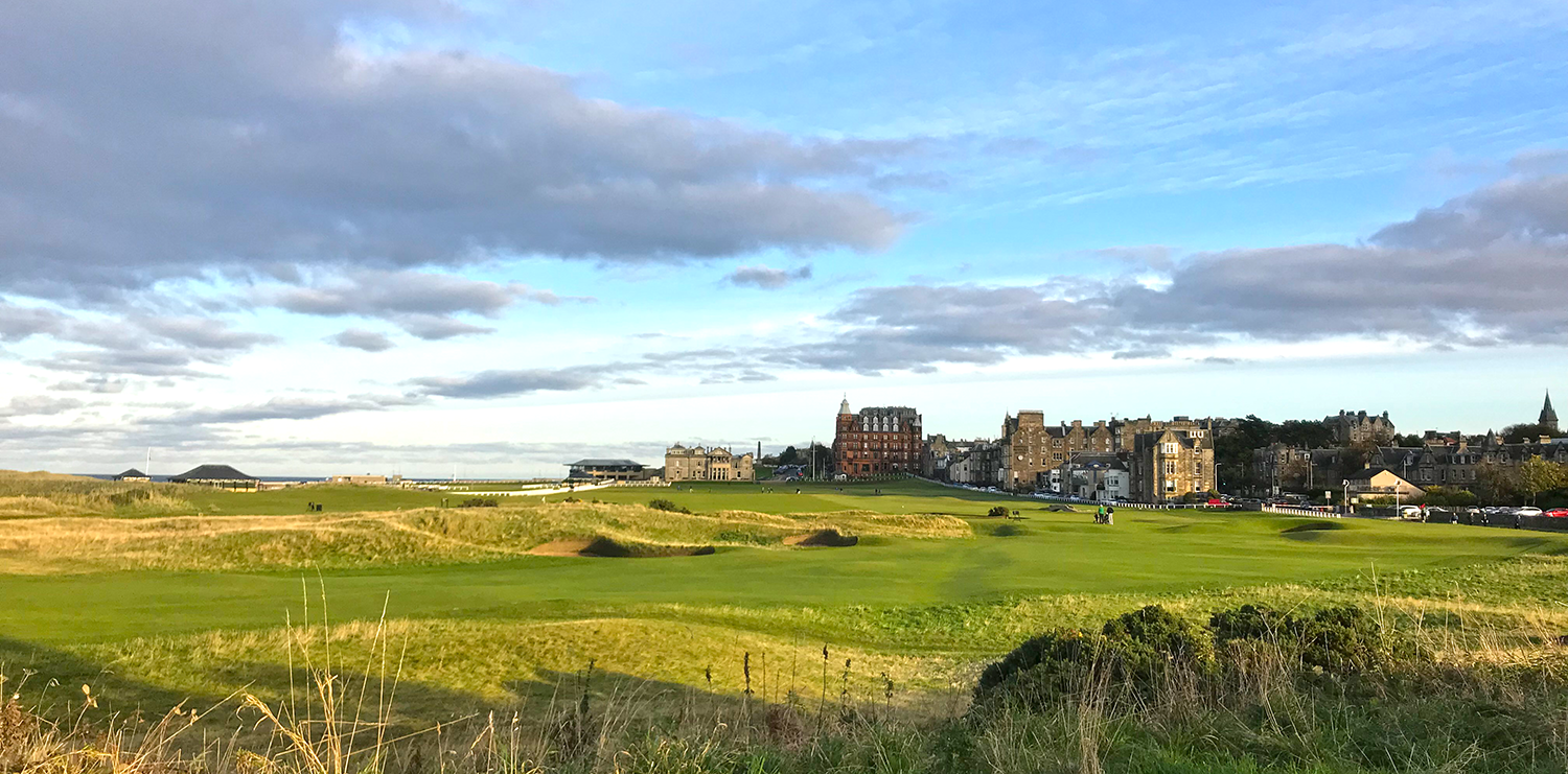 Gröna golfbanor med kullar i förgrunden, historiska stenbyggnader och hotell i bakgrunden under en blå himmel med moln.