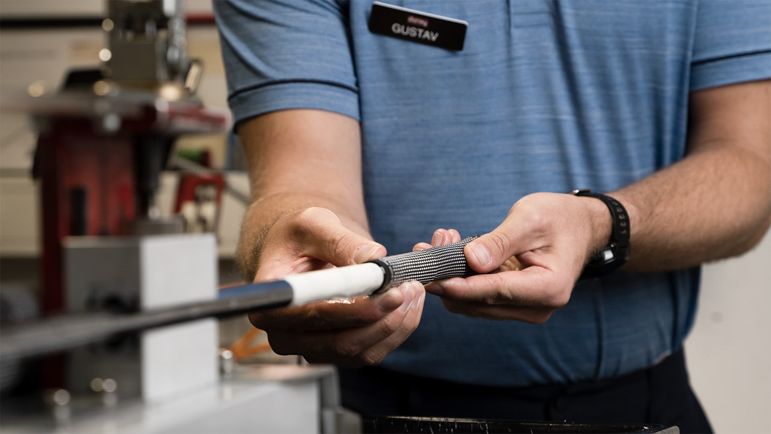 Eine Person mit einem Namensschild „Gustav“ wechselt den Griff eines Golfschlägers in einer Werkstattumgebung.