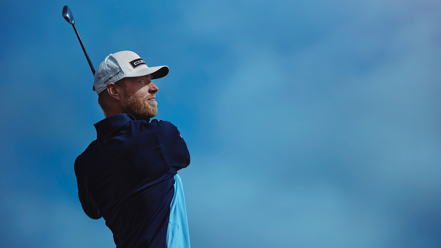 Man svingar en golfklubba utomhus under klarblå himmel, bär vit keps och mörkblå tröja.