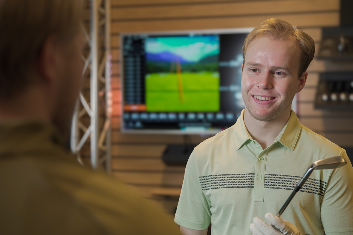 En ung man håller en golfklubba och ler mot en annan person i en golfbutik med en skärm i bakgrunden.