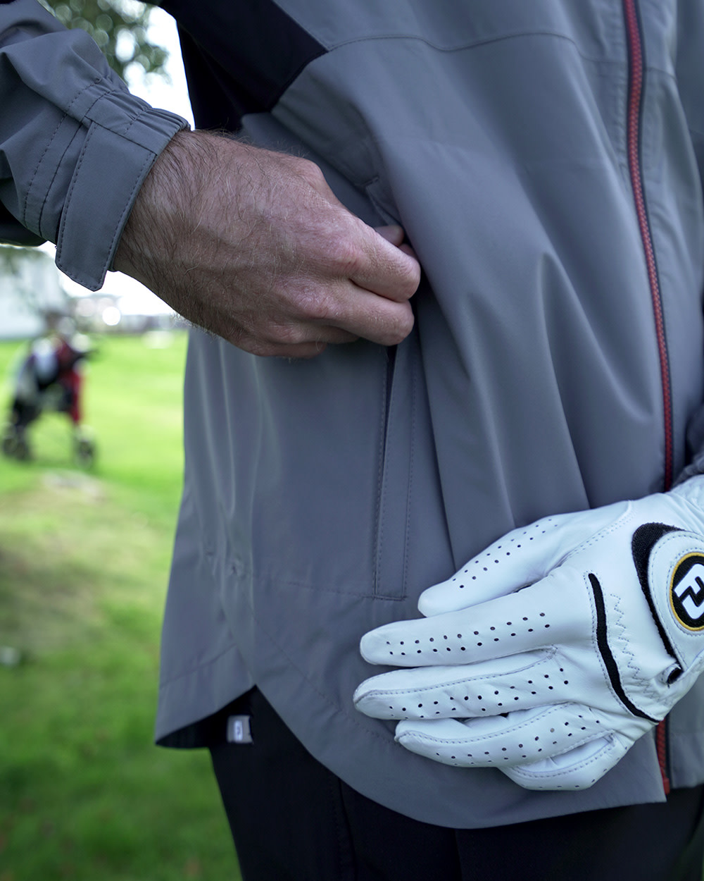 En person i grå jacka stoppar handen i fickan medan den andra handen bär en vit golfhandske, utomhus på en gräsmatta.
