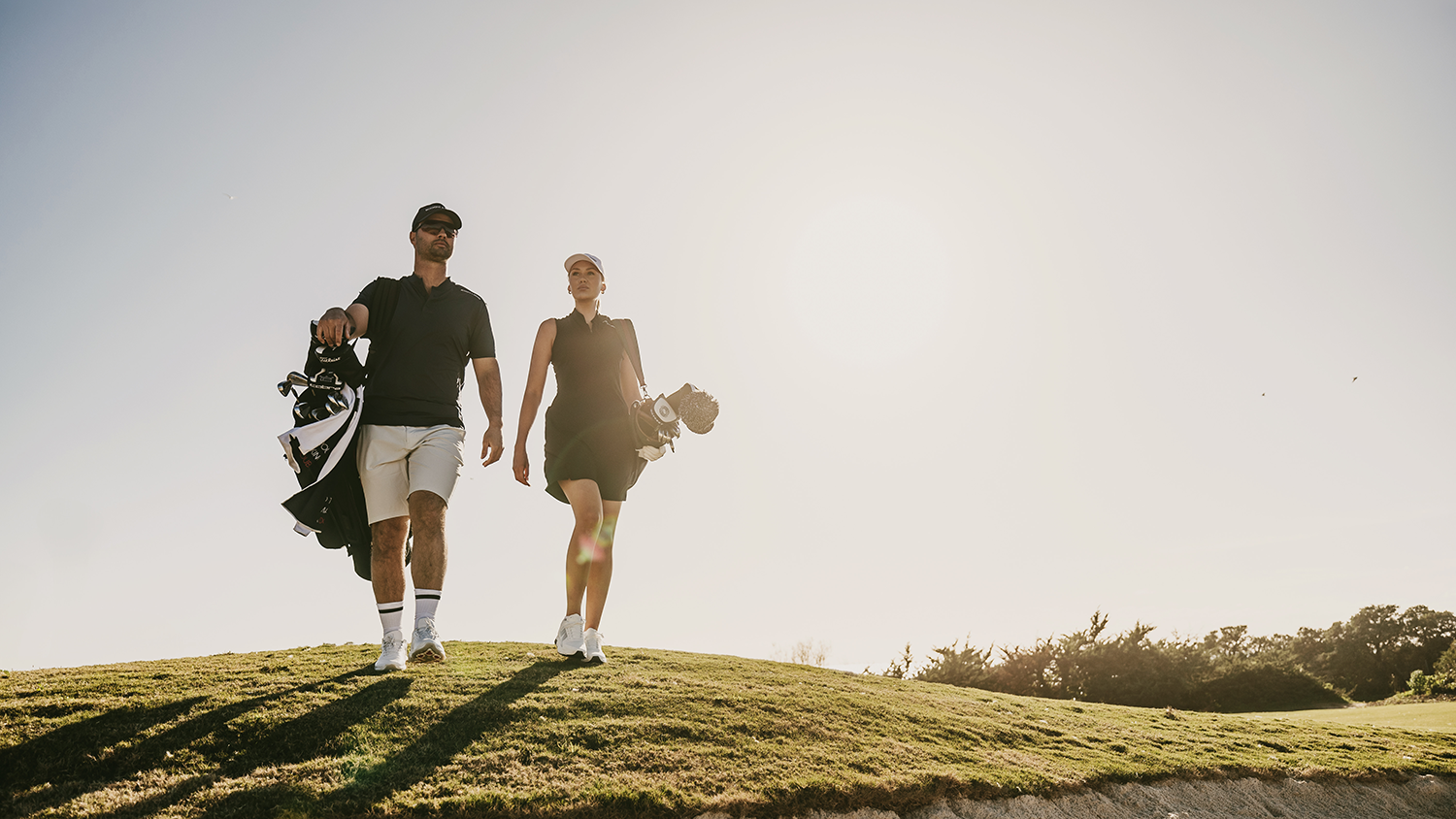 Två golfare går tillsammans på en solig golfbana, bärande på sina golfbagar över axeln.