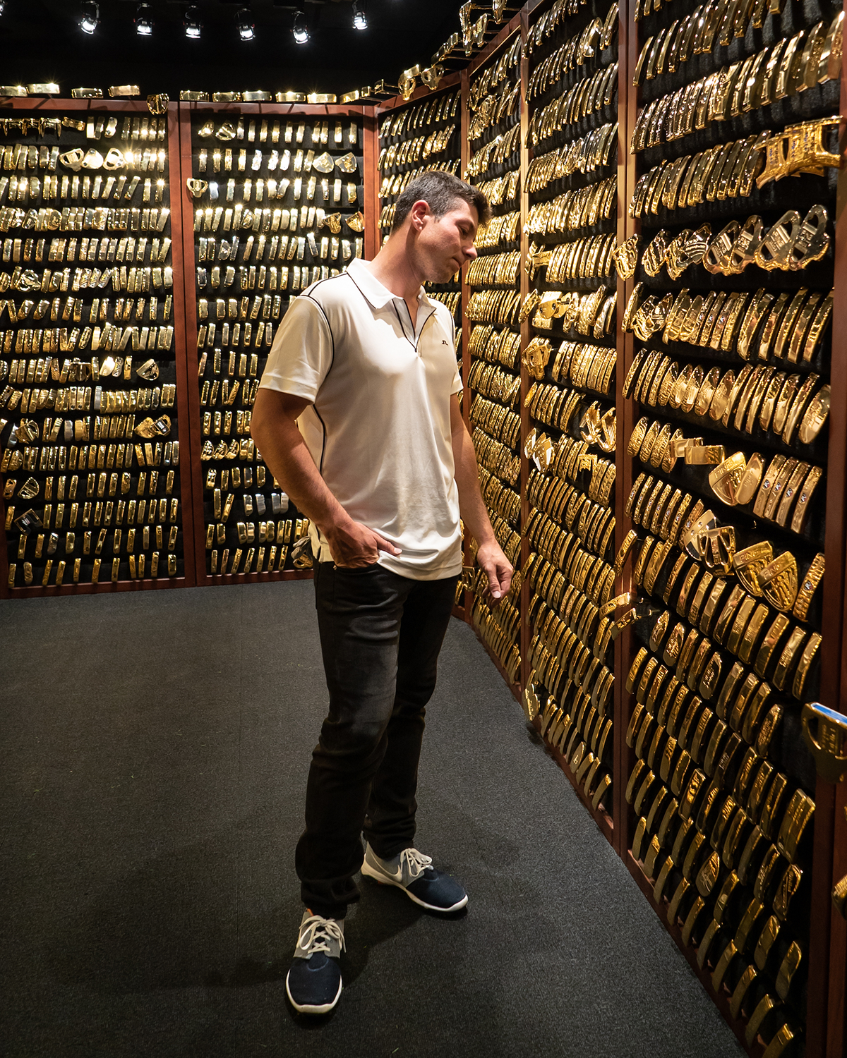 Man i vit pikétröja och svarta byxor tittar på väggar täckta av guldarmband i en butik med mörk belysning.