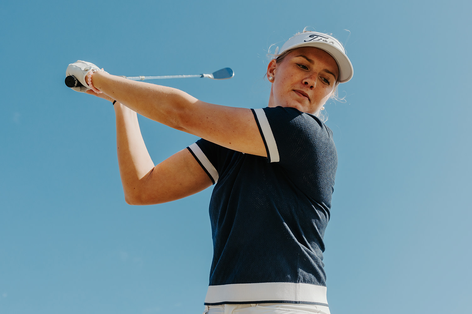 En mann med caps holder en golfkølle bak nakken og ser til siden utendørs i kveldslys.