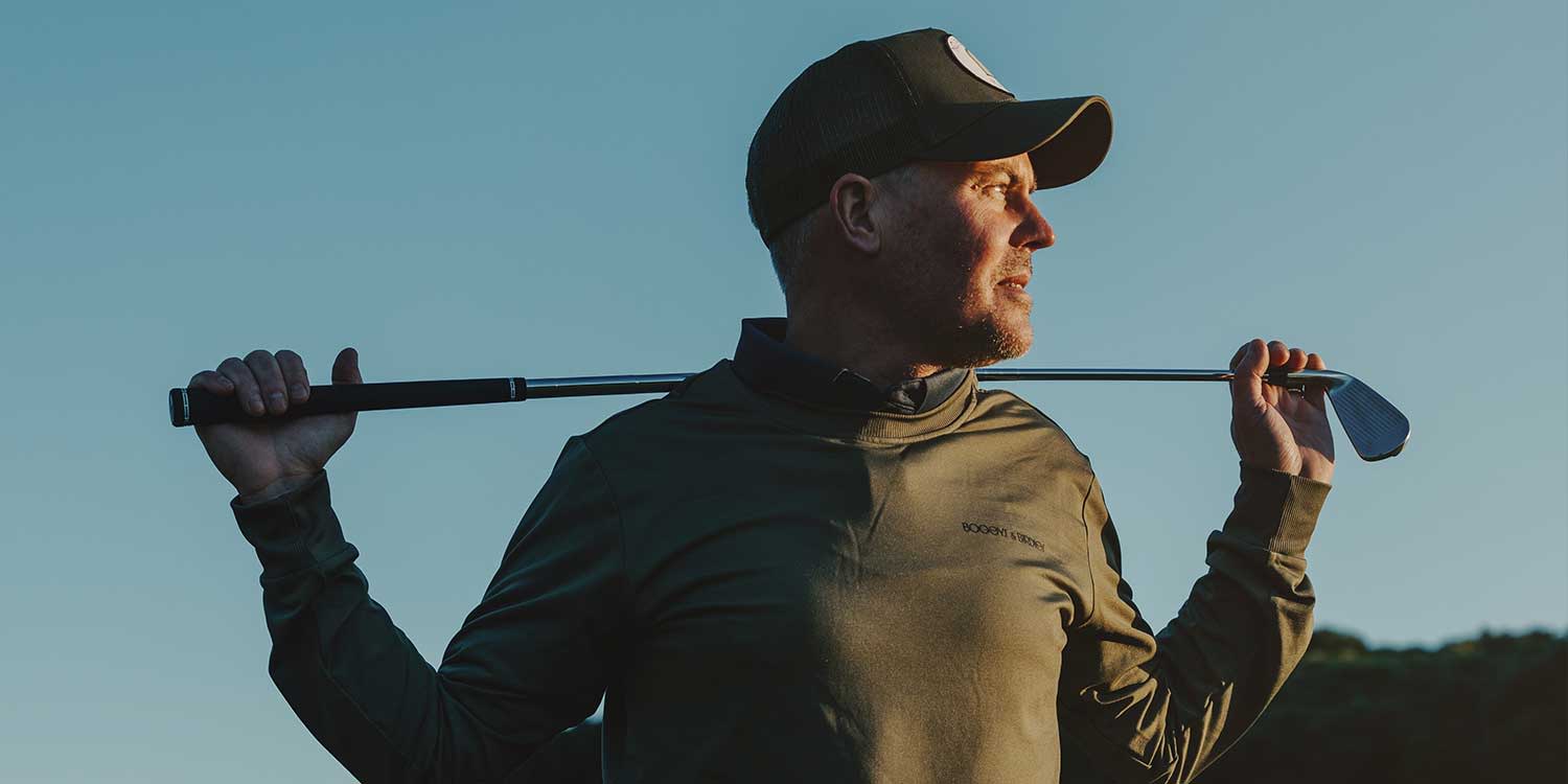 A man in a cap is holding a golf club behind his neck and looking to the side outdoors in the evening light.