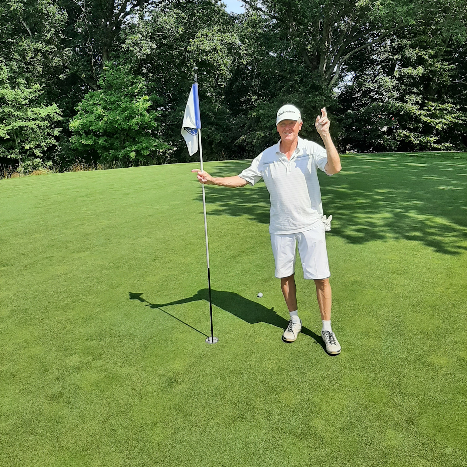 En ældre mand i hvidt golftøj står ved siden af en flagstang på en golfgreen og peger smilende mod hullet.
