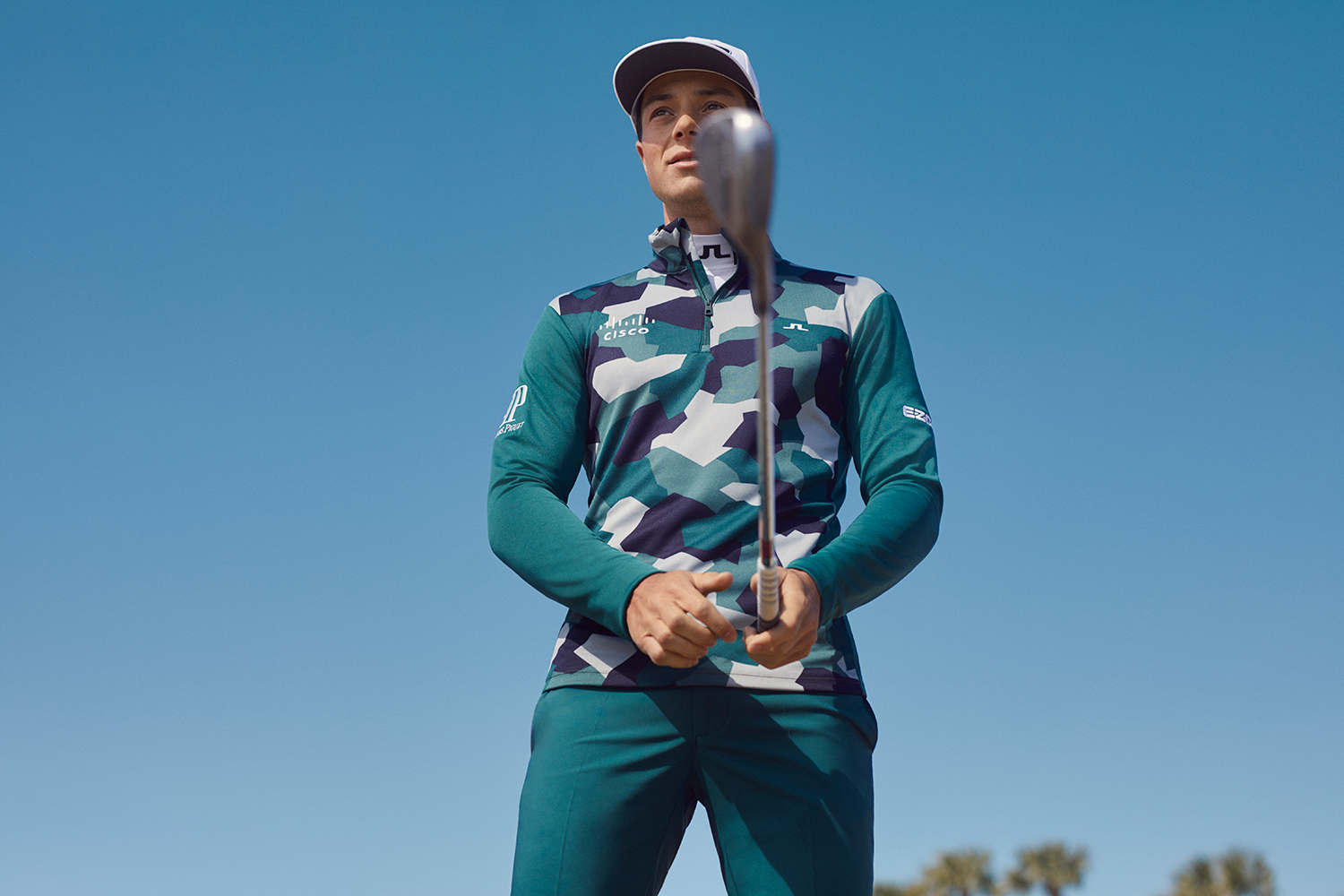 Golfspelare i mönstrad tröja och keps håller en golfklubba mot kameran under klarblå himmel.