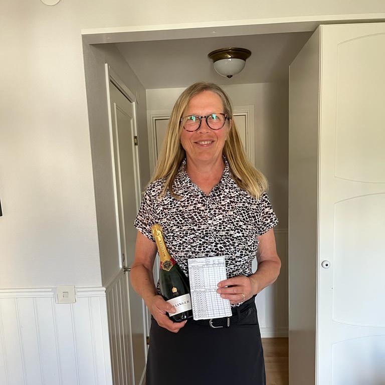 Kvinde med briller holder en flaske mousserende vin og et pointkort, står smilende i en lys entré.