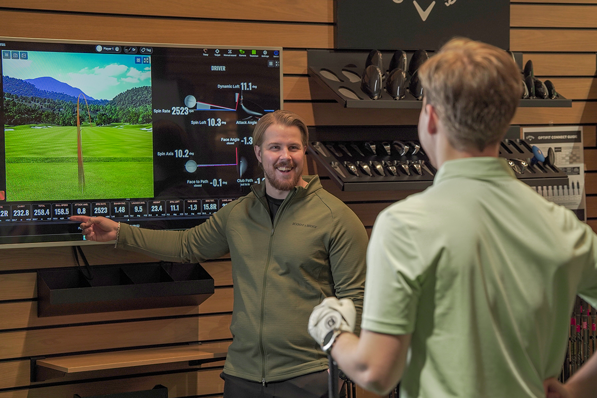 Två män diskuterar golfdata vid en stor skärm i en golfbutik, en pekar på skärmen och den andra håller en golfklubba.