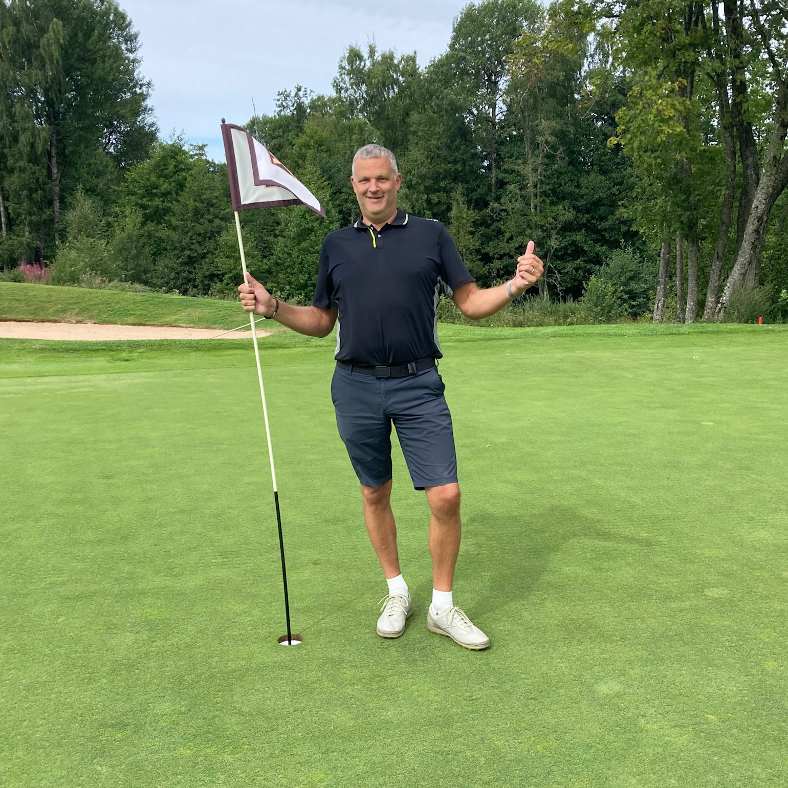Man holder et golfflag og giver tommelfingeren op på en golfgreen med træer i baggrunden.