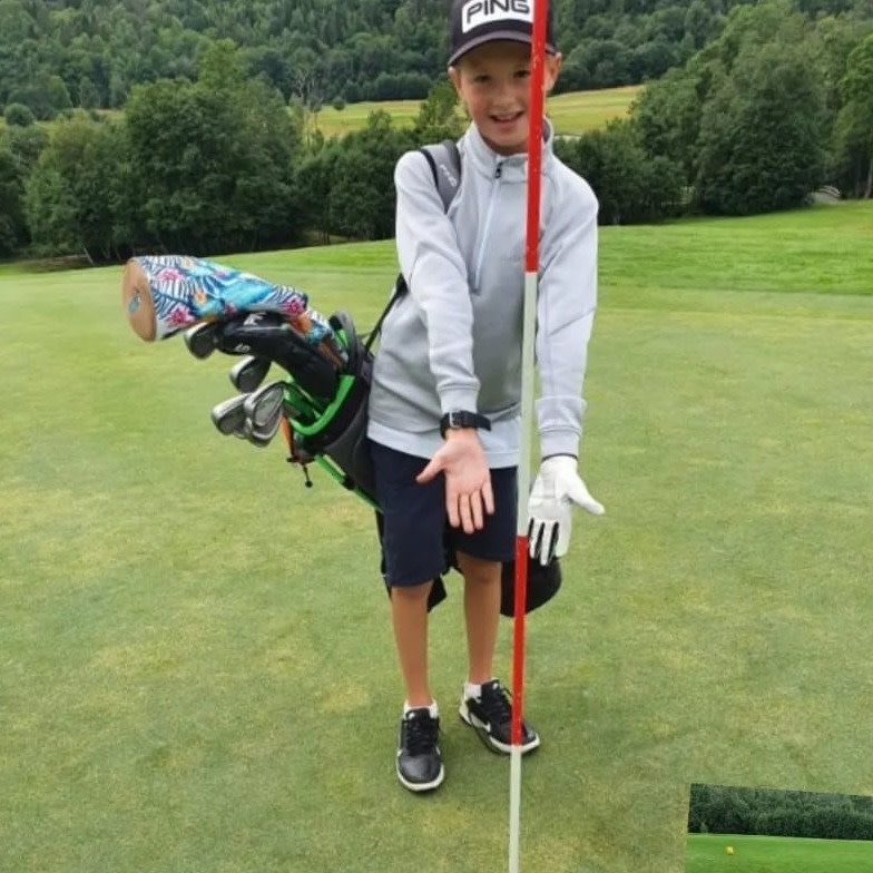Dreng med golfkøller på ryggen står ved siden af en flagstang på en golfbane og peger mod hullet, omgivet af grønne omgivelser.