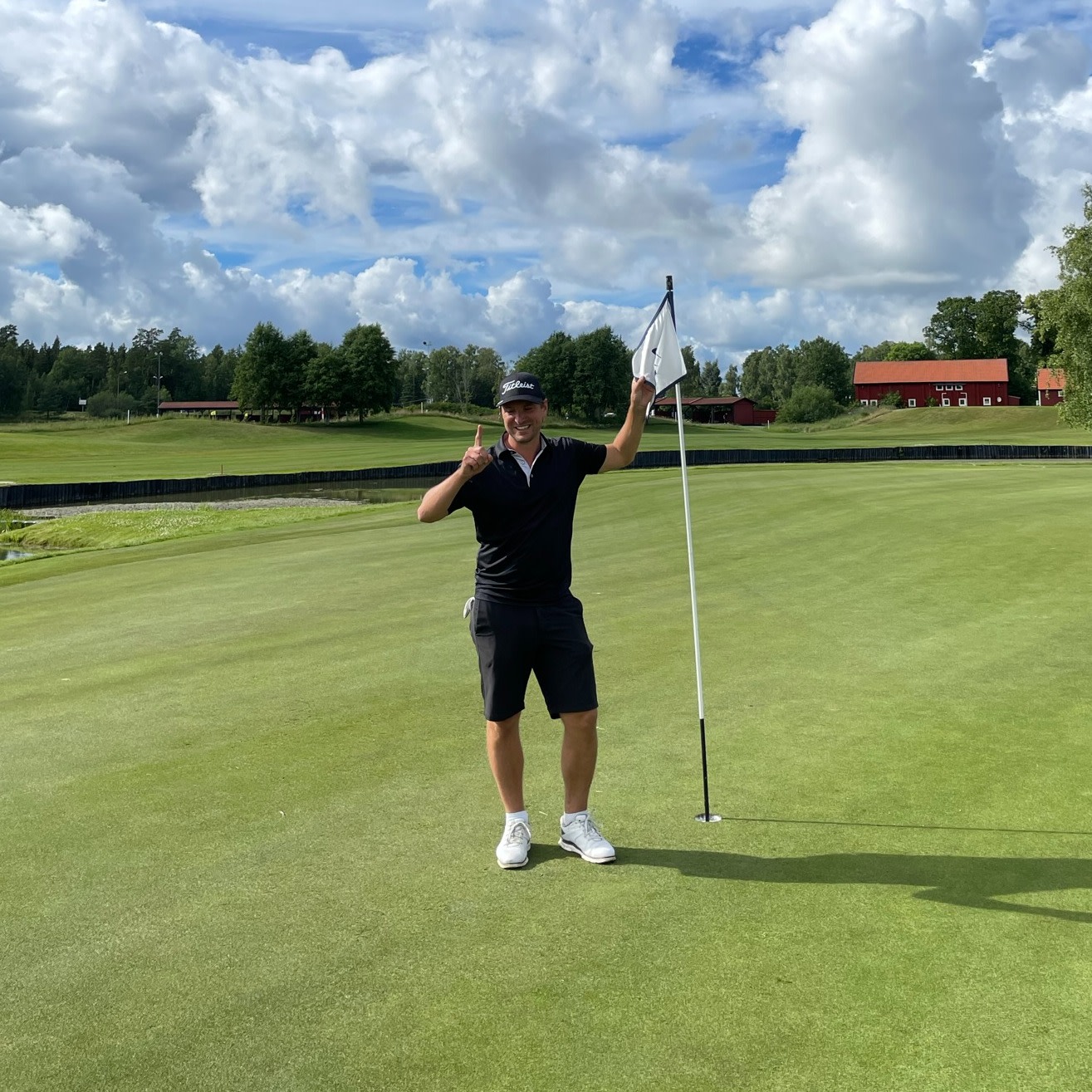 Mand i golfbeklædning står smilende ved en flagstang på en golfbane, holder én finger op, grønt landskab og røde huse i baggrunden.