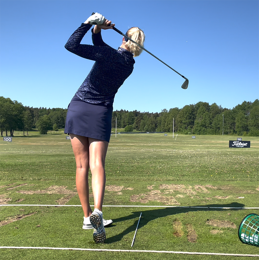 Kvinna svingar en golfklubba på en driving range, omgiven av grönt gräs och träd under en klarblå himmel.