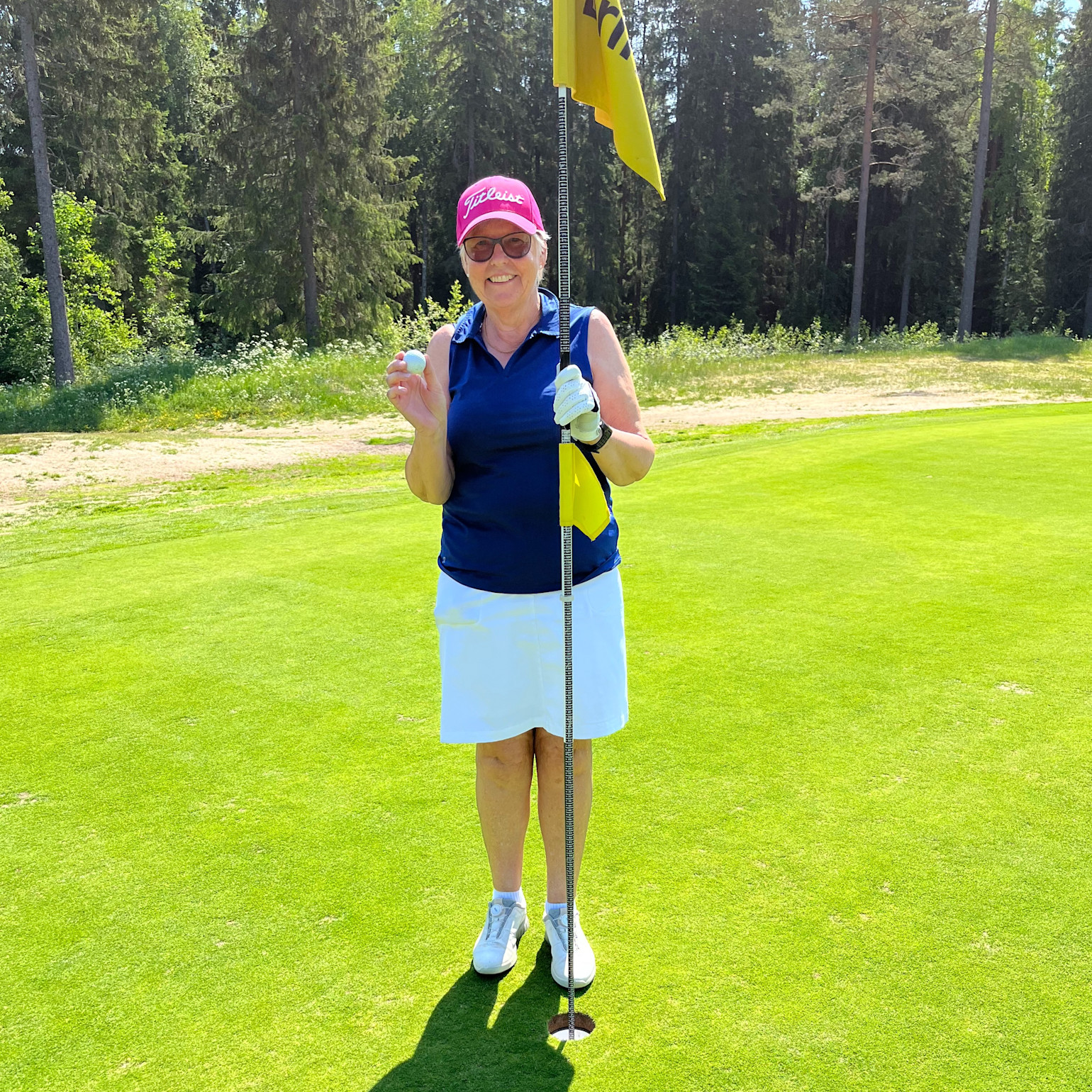 Kvinde med lyserød kasket og solbriller holder en golfbold og står ved flagstangen på en golfgreen, smilende i solskinnet.