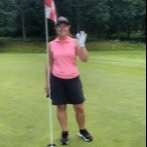 Kvinde i lyserød trøje og sort nederdel står på en golfbane, holder i en flagstang og vinker mod kameraet.