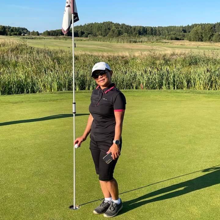 Kvinde i sort golfbeklædning står ved flagstangen på en golfgreen, holder en golfbold og en mobiltelefon, solrigt landskab i baggrunden.