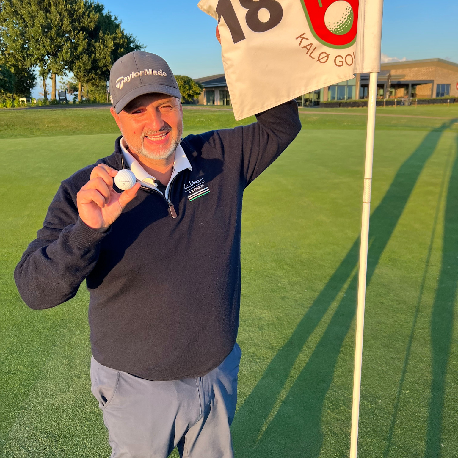 Mand iført golfbeklædning holder en golfbold og et flag ved hul 18 på en golfbane i solskinsvejr.
