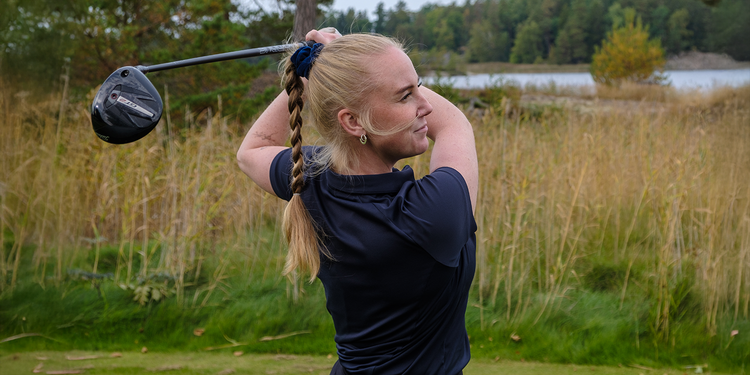Frau mit Zopf schwingt einen Golfschläger auf einem Golfplatz mit hohem Gras und Bäumen im Hintergrund.