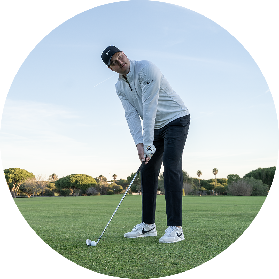 Mies urheiluvaatteissa valmistautuu lyömään golfpalloa vihreällä golfkentällä, taustalla puita.