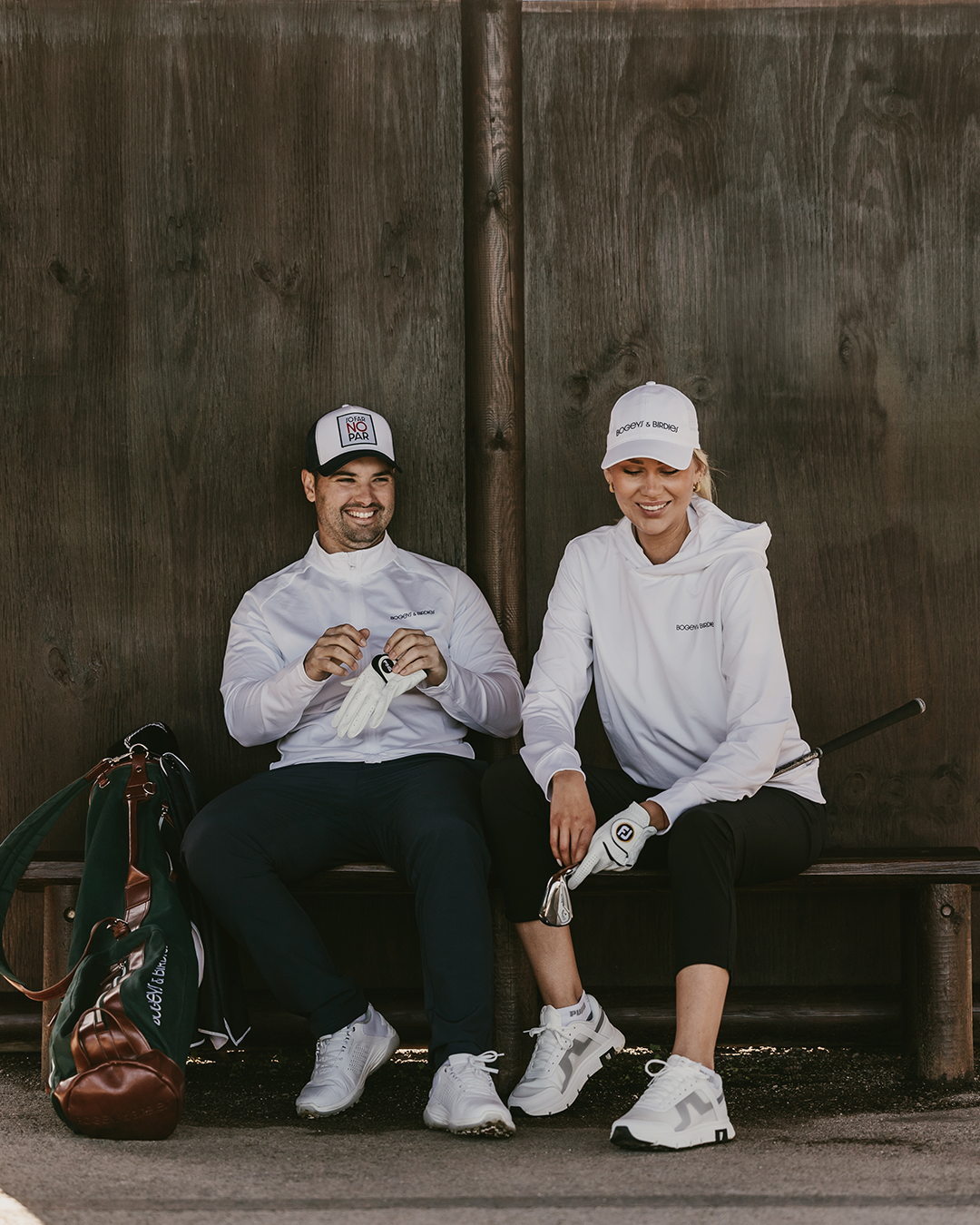 Två golfare i vita tröjor och kepsar sitter på en bänk, skrattar och förbereder sig med golfhandskar, golfbag bredvid.