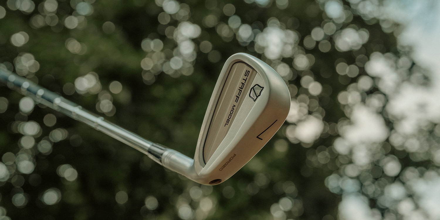 En golfkølle med teksten "STAFF MODEL" holdes opp mot en uklar bakgrunn av grønne trær og lyse prikker.