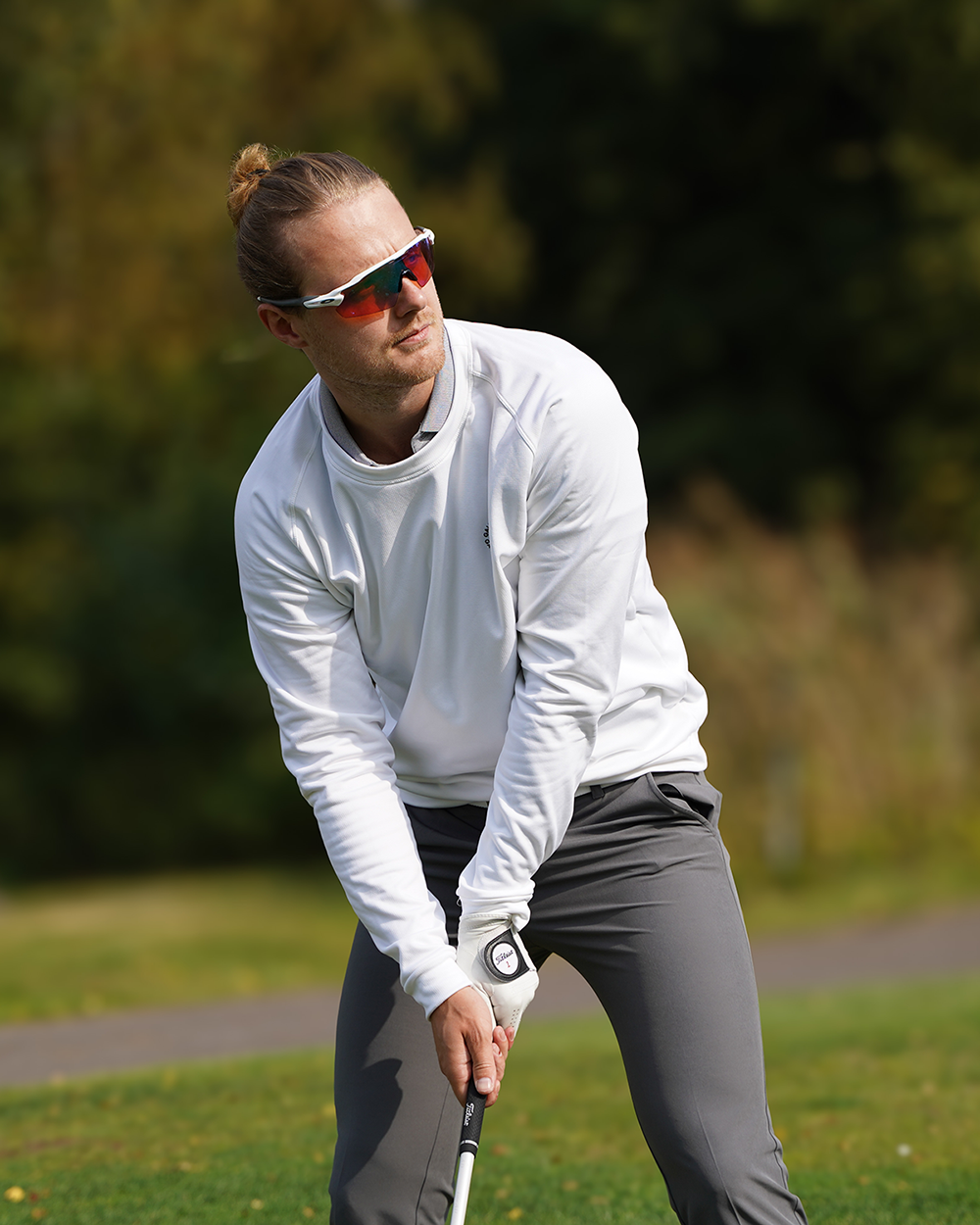 Mies, jolla on aurinkolasit ja valkoinen paita, valmistautuu lyömään golfpalloa vihreällä ulkona olevalla golfkentällä.