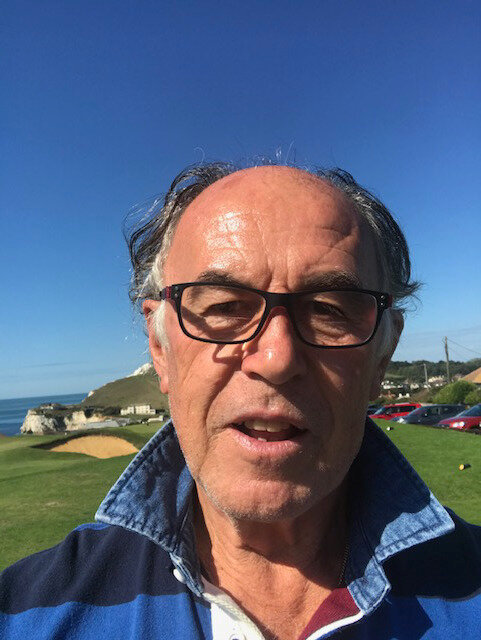 Man med glasögon står utomhus på en golfbana med grönt gräs, blå himmel och kullar i bakgrunden.