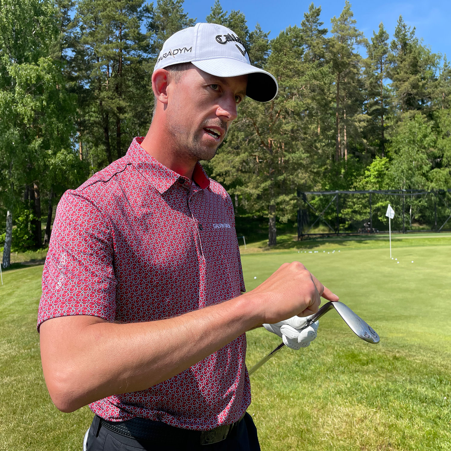 Man i röd mönstrad golftröja och vit keps håller en golfklubba och pratar vid en green på golfbana med träd i bakgrunden.