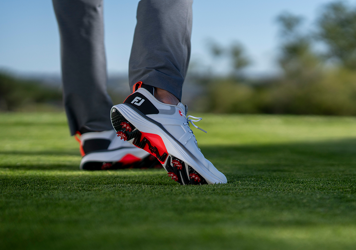 Person går på gräs med grå byxor och vita golfskor med svarta och röda detaljer, logotypen "FJ" synlig på hälen.