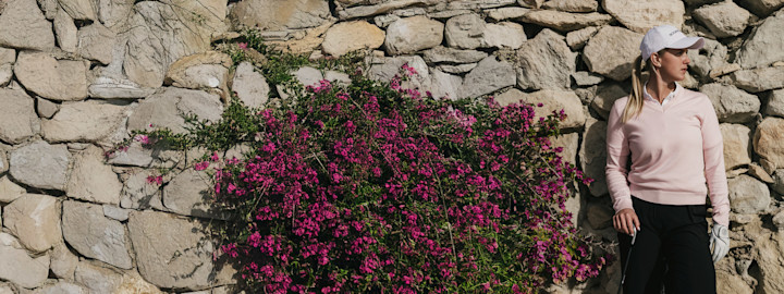 Kvinna i blå tröja svingar en golfklubba utomhus under blå himmel med träd i bakgrunden.