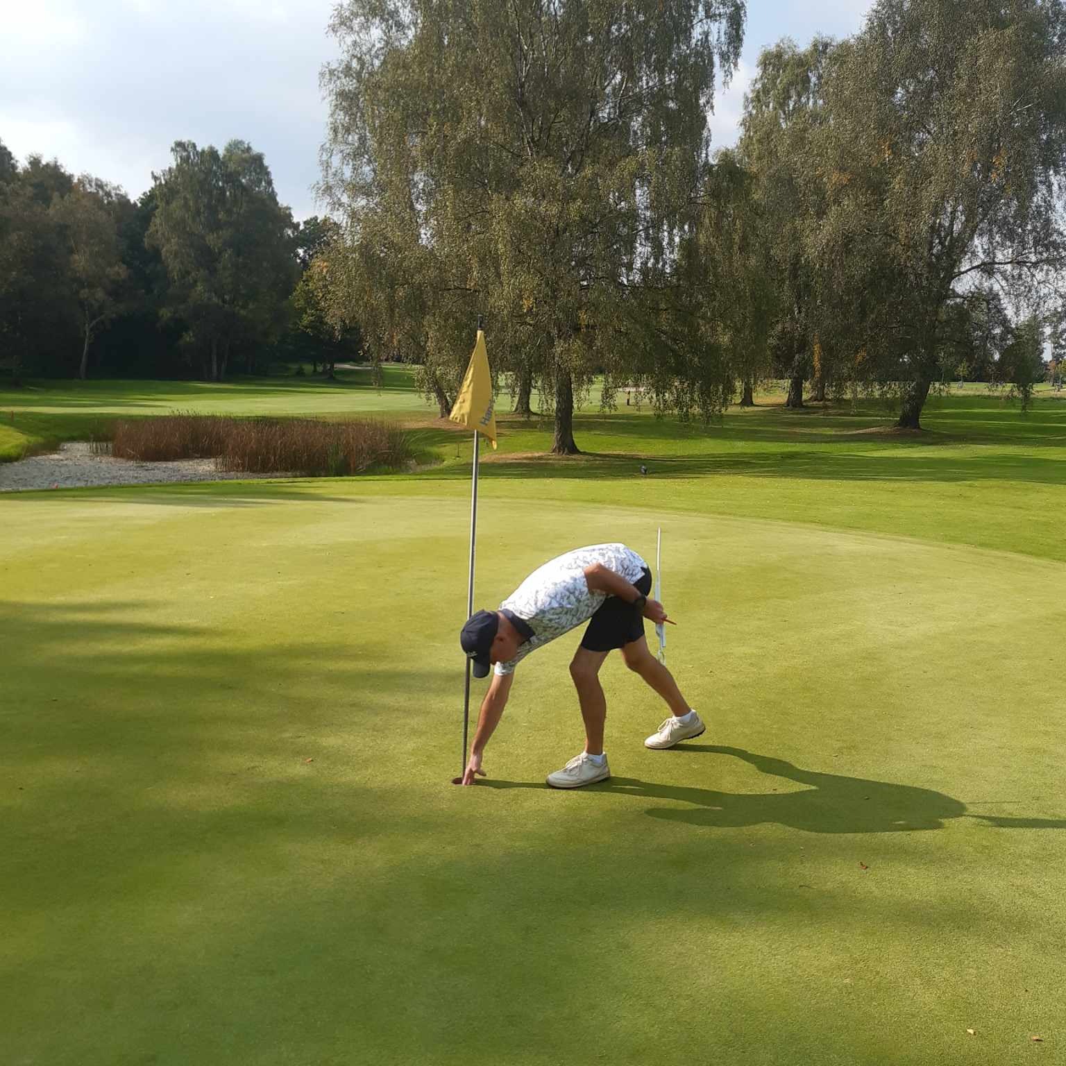 En golfspiller bøjer sig ned for at samle en golfbold op ved hullet på en grøn golfbane omgivet af træer.
