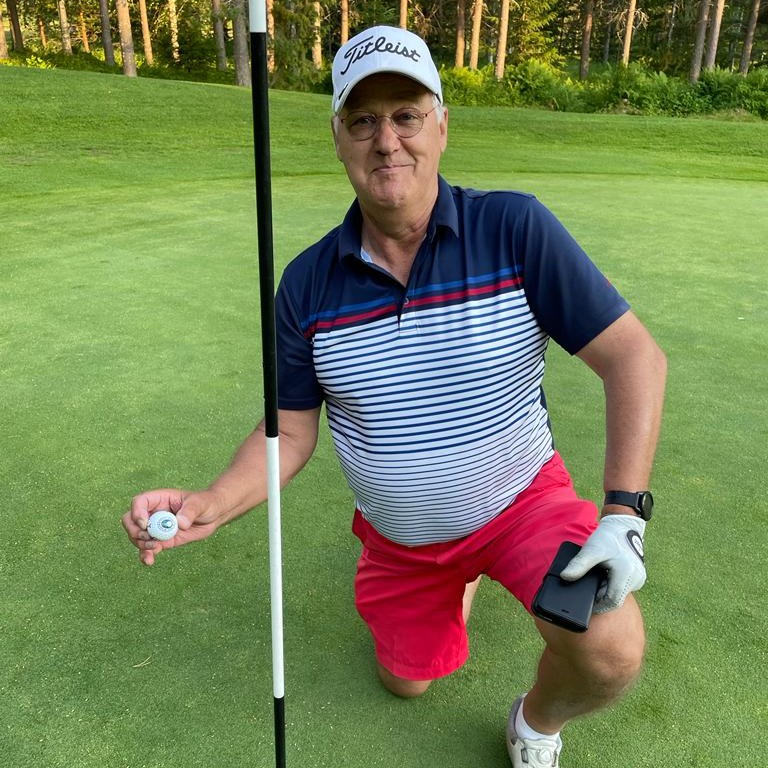 Mand på en golfgreen iført en stribet polo og røde shorts sidder på hug, holder en golfbold ved siden af hullet og smiler til kameraet.