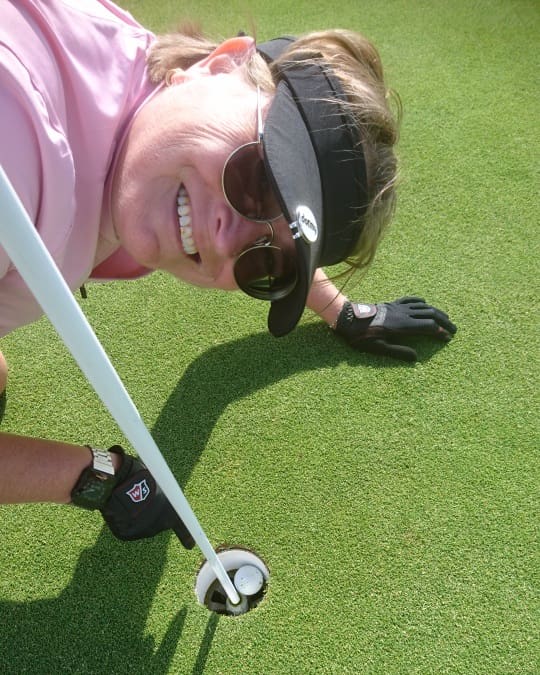 Person med solbriller og visir smiler ved siden af et golfhul, hvor en golfbold ligger på greenen.