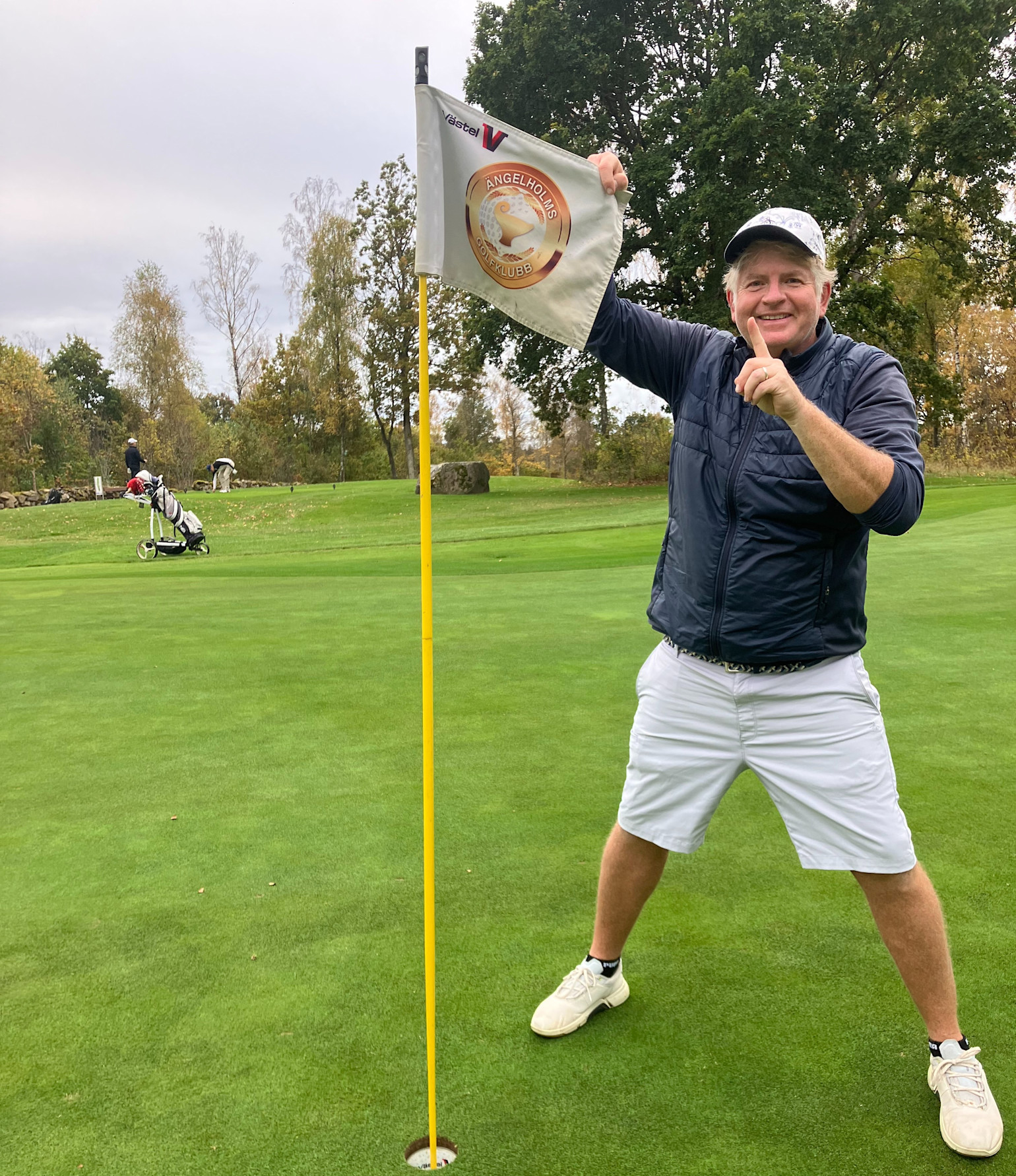En mann i golfklær står ved siden av en flaggstang på greenen, holder i flagget med teksten "Ängelholms Golfklubb" og smiler mot kameraet.