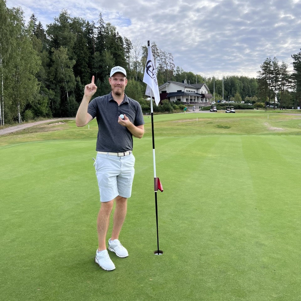 Mand iført golfbeklædning holder en finger og en golfbold op ved siden af et hul med flag på en golfbane, med klubhus i baggrunden.