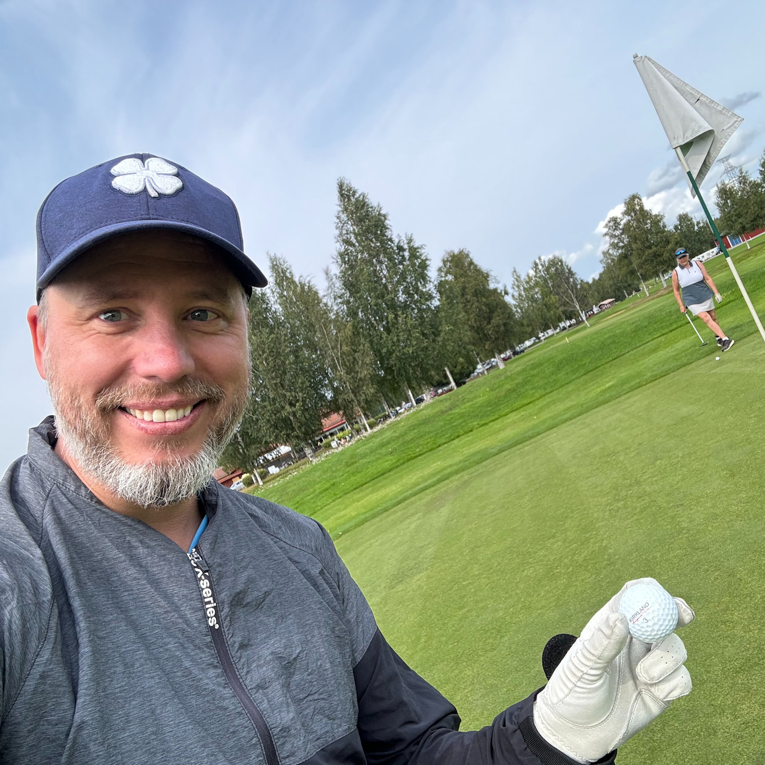 Hymyilevä mies, jolla on lippis ja golfhanska, pitää golfpalloa greenillä. Taustalla näkyy lippukeppi ja toinen golfari.