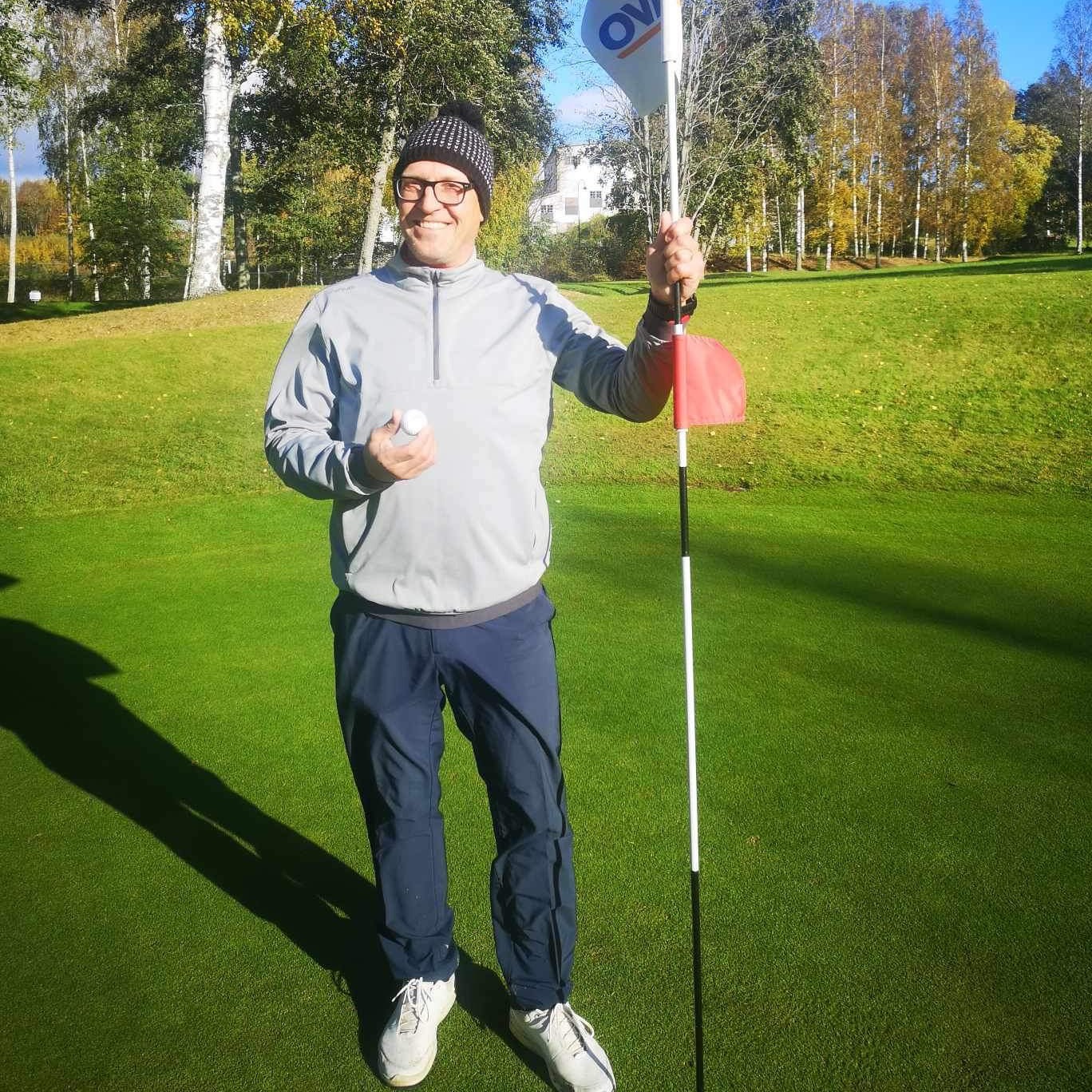 Man i mössa och glasögon håller en golfboll och flaggstång på en golfbana med grönt gräs och träd i bakgrunden.