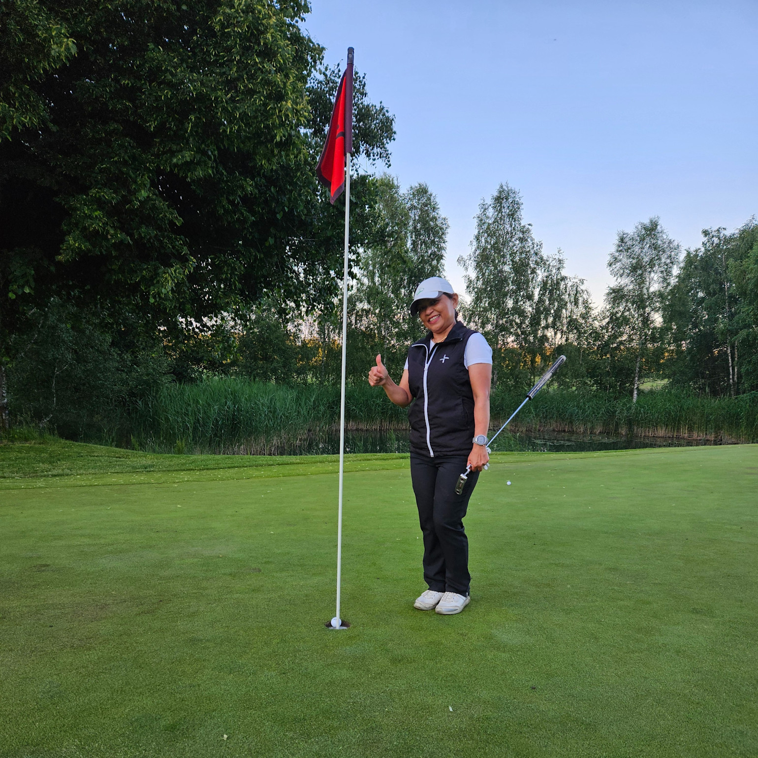 Kvinde med golfkølle giver tommelfingeren op ved siden af et golfhul med flag på greenen, omgivet af træer og græs.