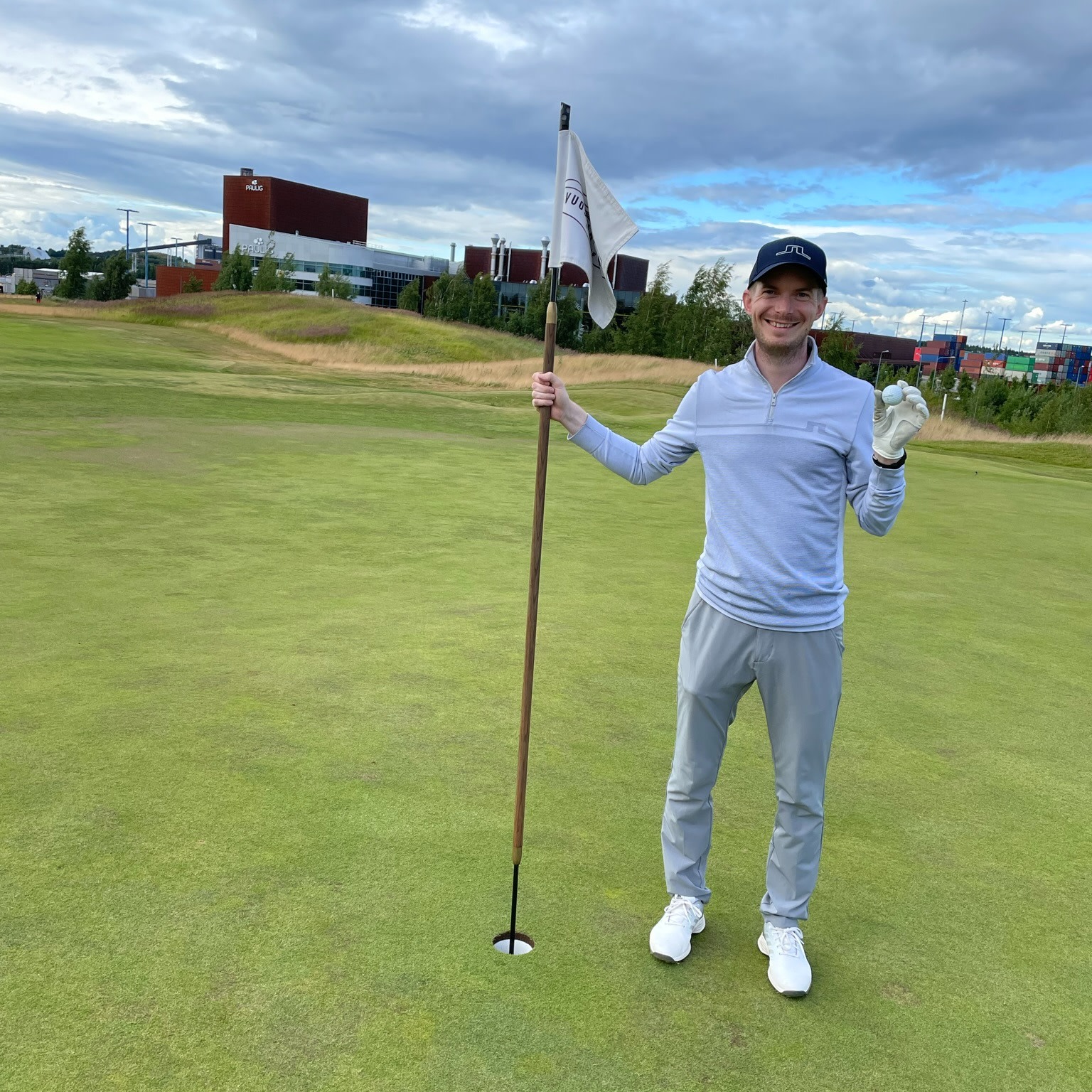 Man holder en flagstang ved et golfhul og viser en golfbold frem på en golfbane med bygninger i baggrunden.