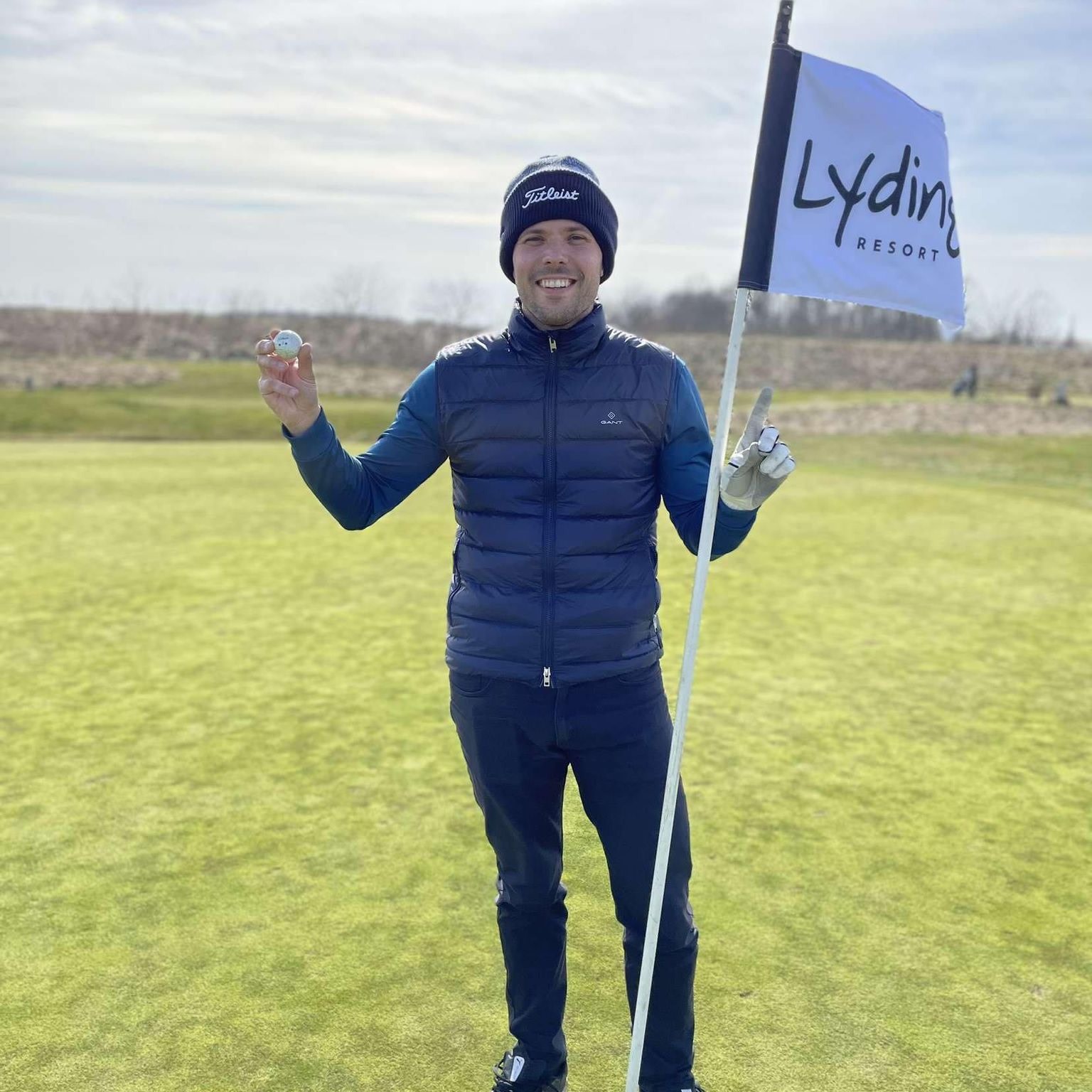 A person is holding a golf ball and standing next to a flagpole with the text "Lydinge Resort" on the golf course.