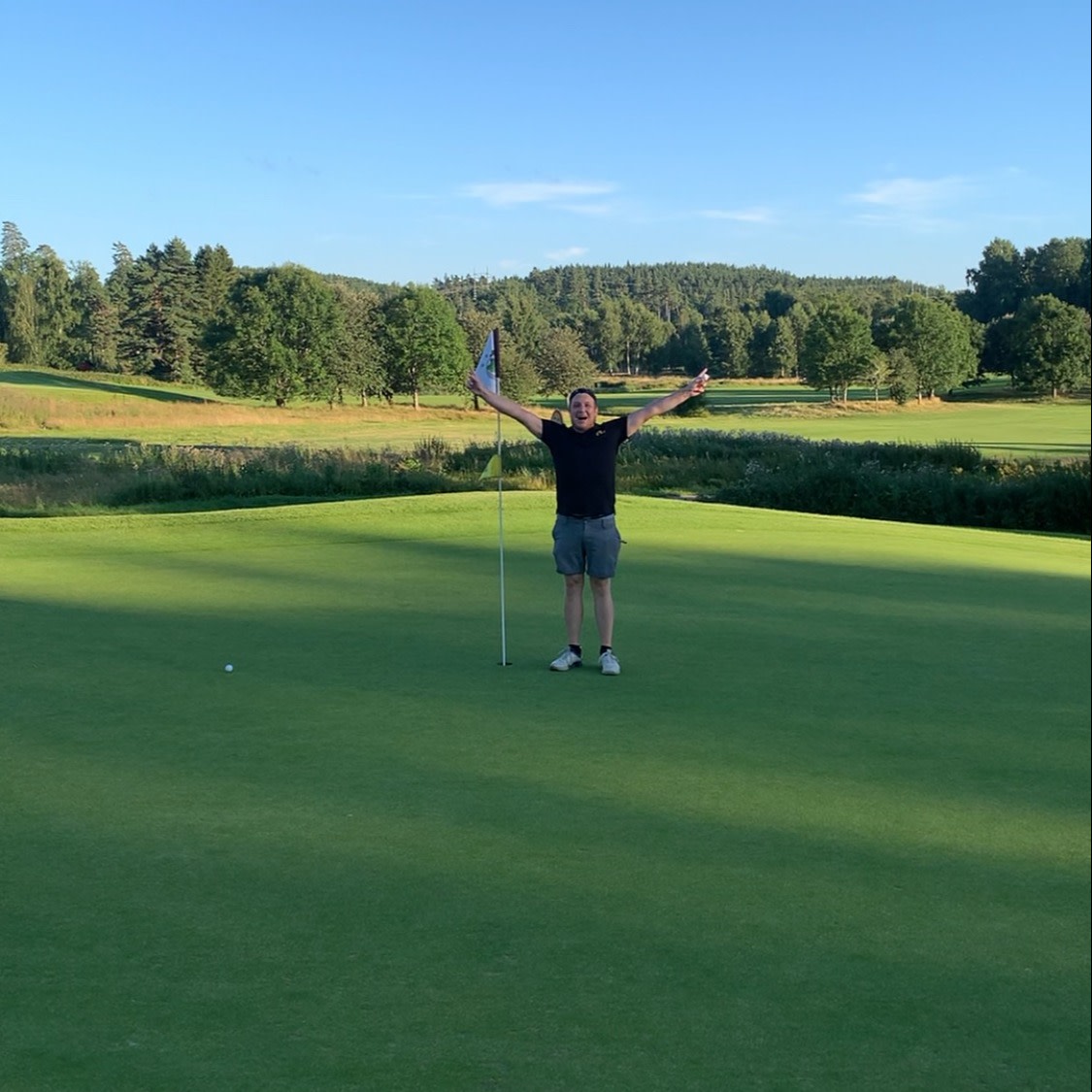 En mand fejrer med armene i vejret ved siden af en flagstang på en golfgreen, omgivet af grønne områder og træer under en klar himmel.