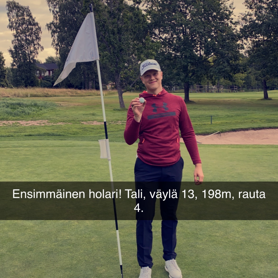 En ung mand i rød trøje og kasket holder en golfbold op ved siden af en flagstang på en green, med træer i baggrunden.