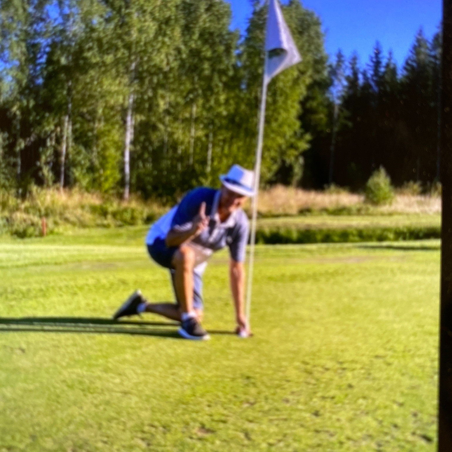 Mand iført hat poserer og laver sejrtegn ved et golfhulflag på en grøn golfbane med træer i baggrunden.