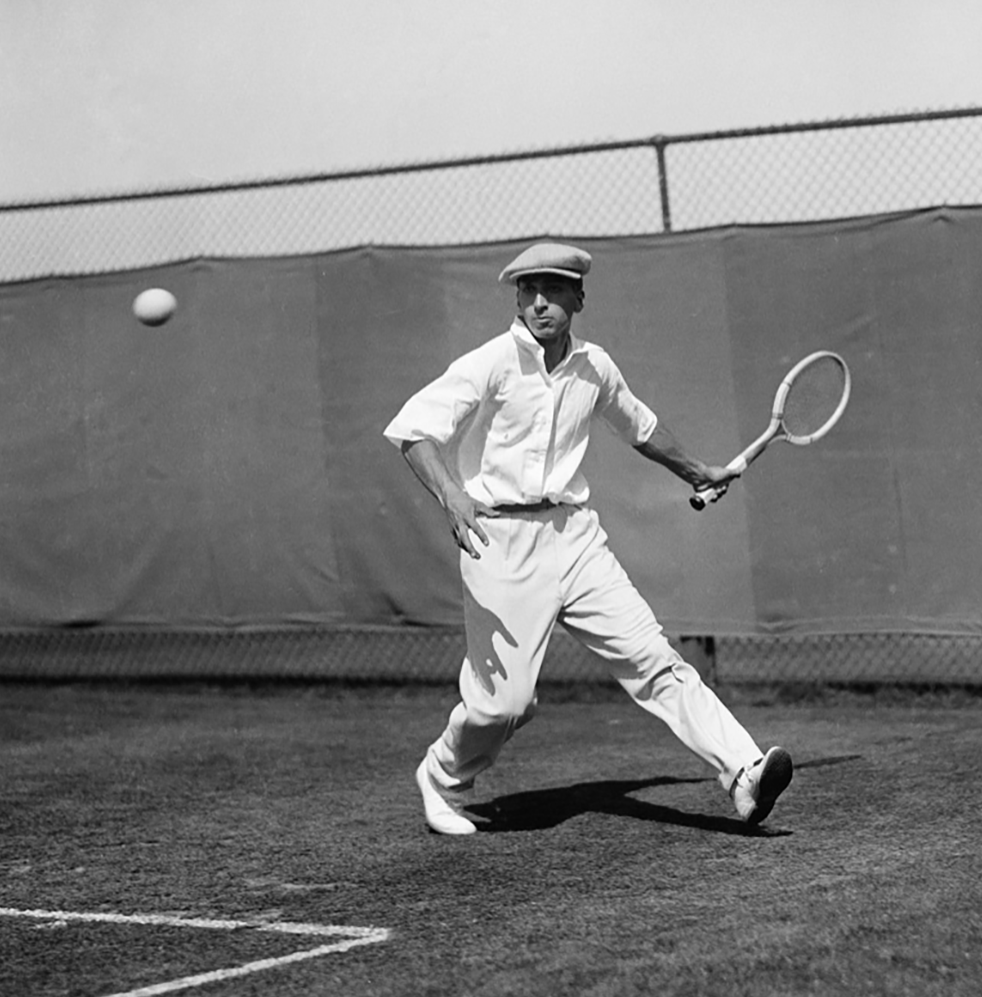 Manlig tennisspelare i vit klädsel och keps slår en boll med racket på en gräsbana, stängsel i bakgrunden.