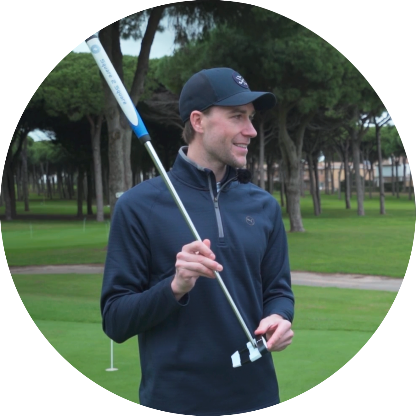 A man in a dark cap and jumper is holding a golf club on a golf course with green grass and trees in the background.